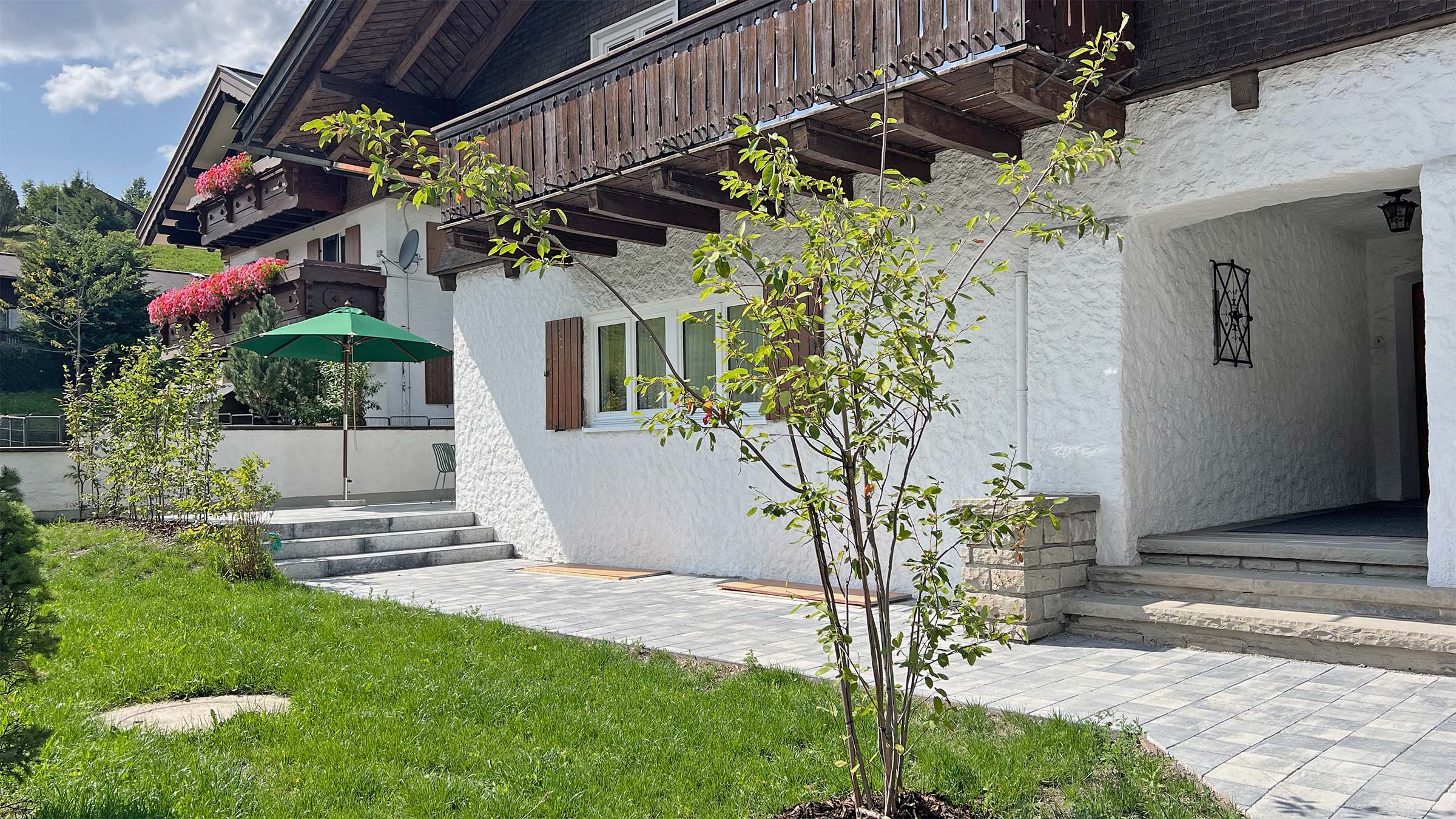 Der Hauseingang der Talstation im Sommer, davor Rasen und ein Sonnenschirm auf dem Balkon