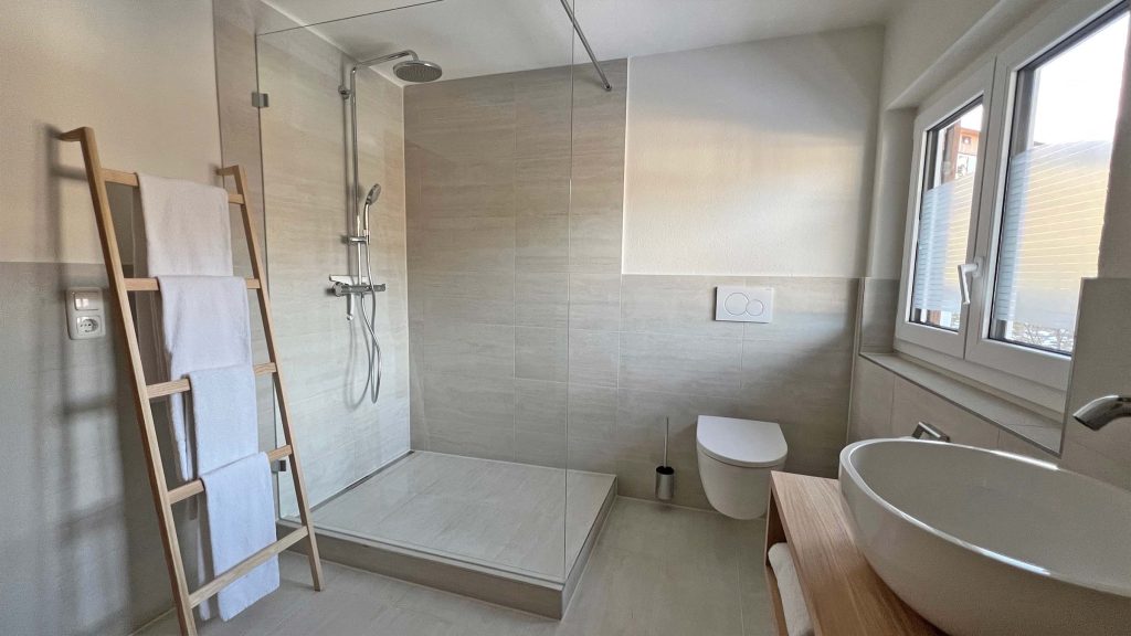 Bodengleiche Dusche hinter Glas, daneben WC und Handtuchleiter